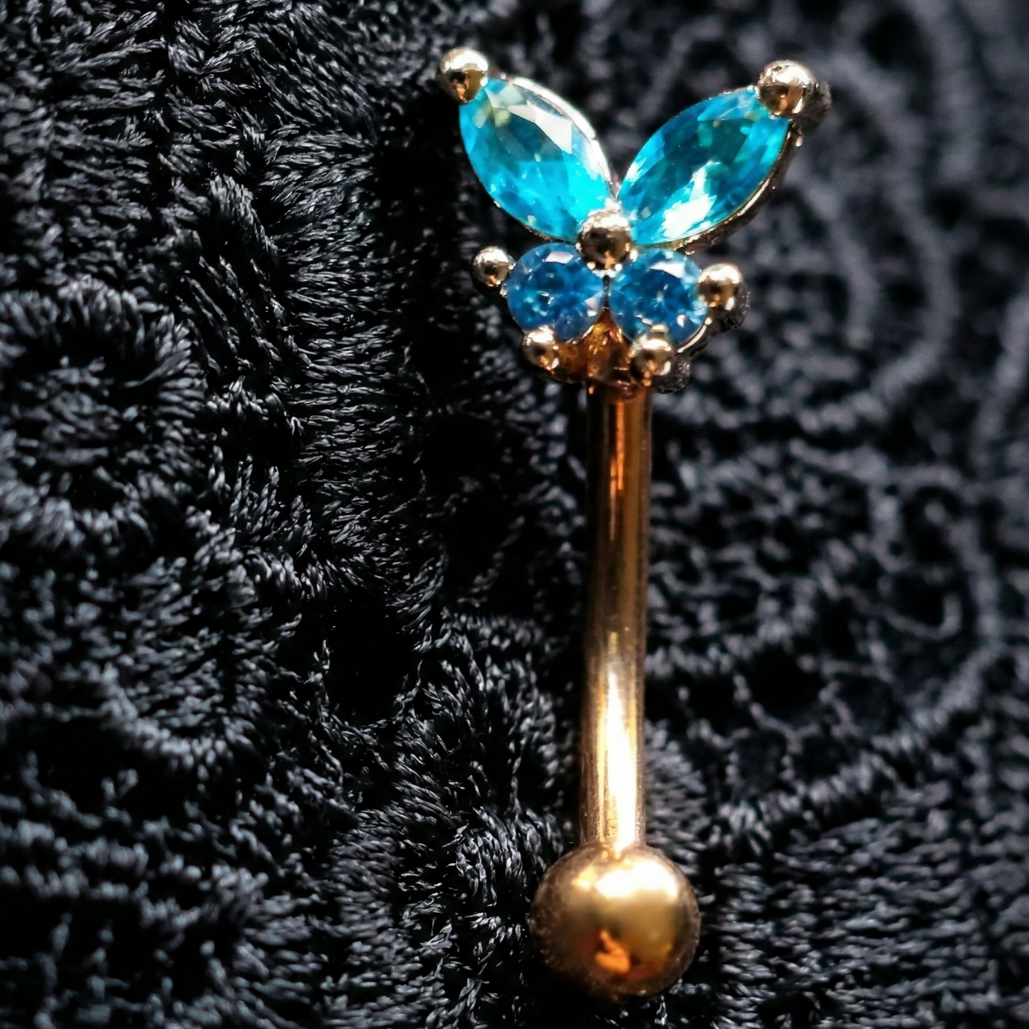 Butterfly-shaped eyebrow piercing jewellery with blue gemstones on a textured black background