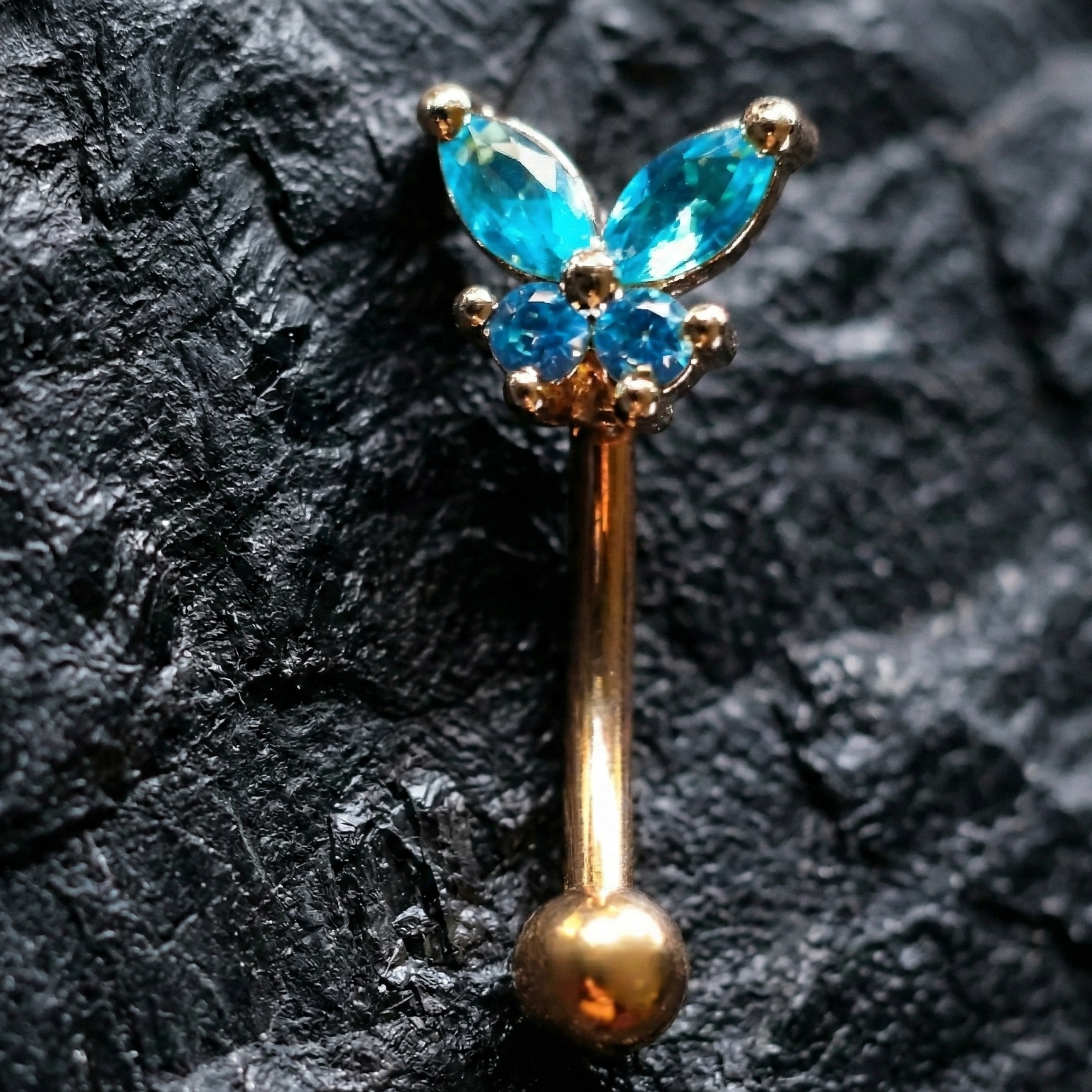 Butterfly-shaped eyebrow/rook barbell with blue gemstones on a dark textured background