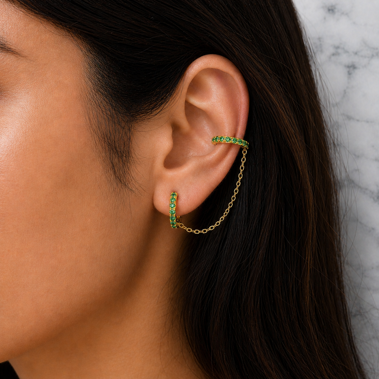 Close-up of an ear wearing a gold hoop earring with green gemstones against a marble background