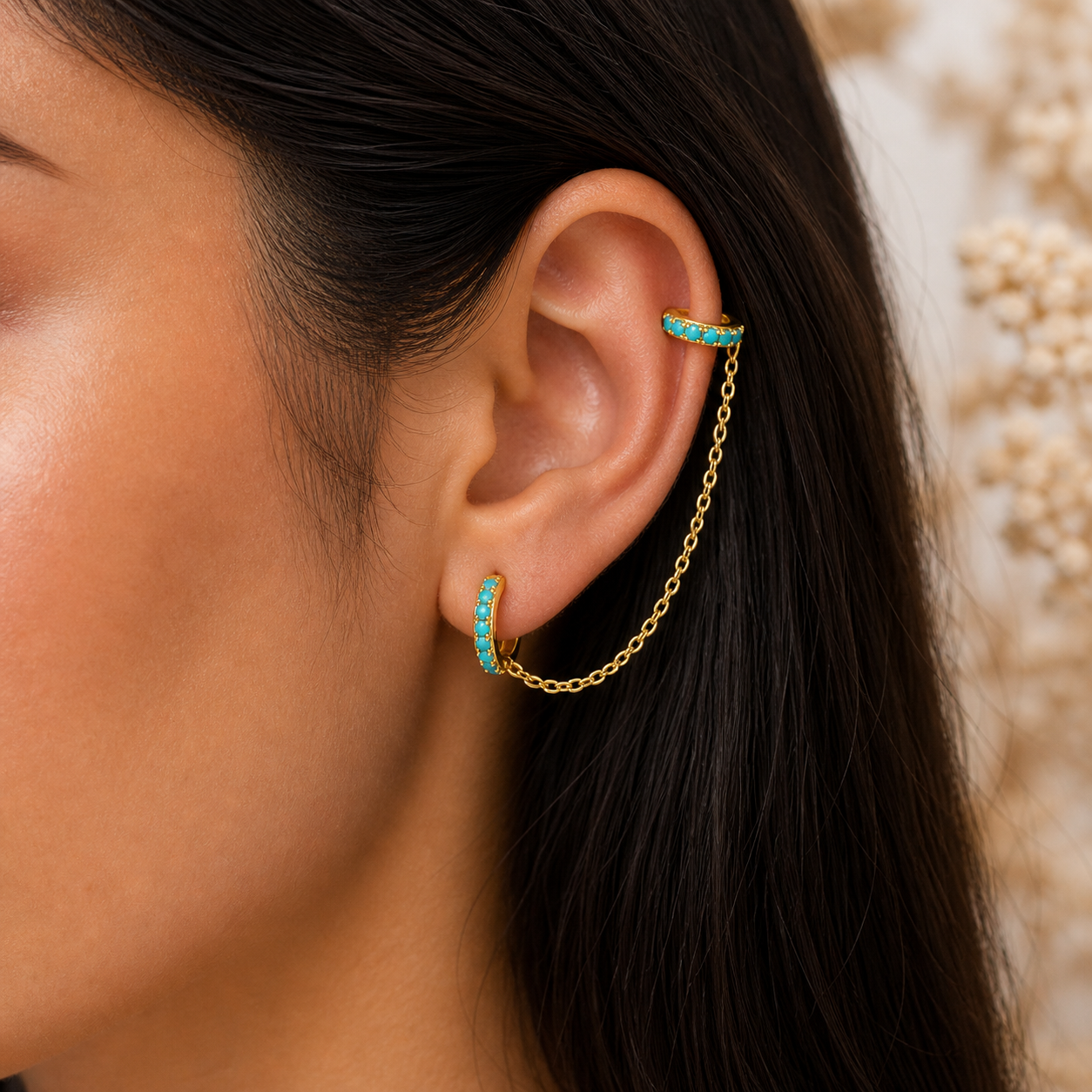 Close-up of an ear with turquoise earrings and a gold chain earring against a neutral background