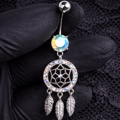 Belly button ring with dreamcatcher design held by a gloved hand on a textured black background
