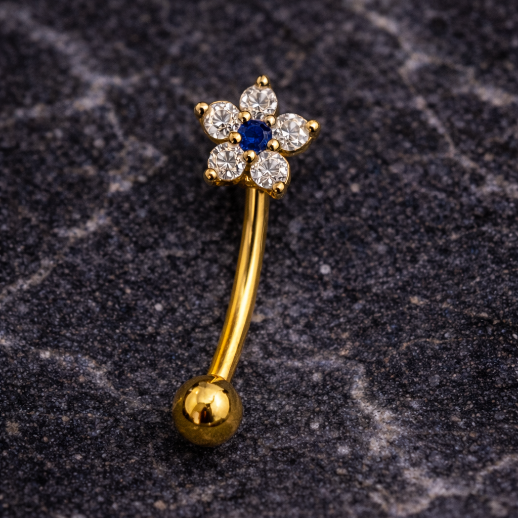 Gold eyebrow/rook barbell with a flower design on a dark textured surface