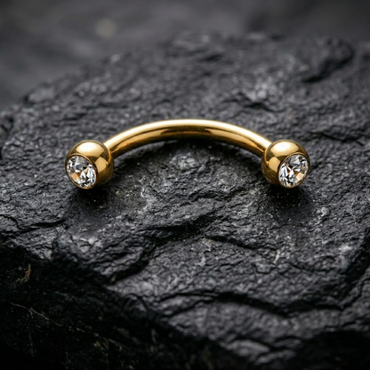Gold eyebrow/rook barbell with crystals on a dark stone surface