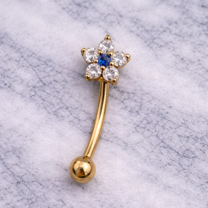 Gold eyebrow/rook piercing bar with star-shaped top and blue gemstone on a marble background