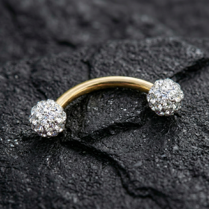 Gold eyebrow/rook piercing ring with two diamond-like spherical ends on a dark textured surface