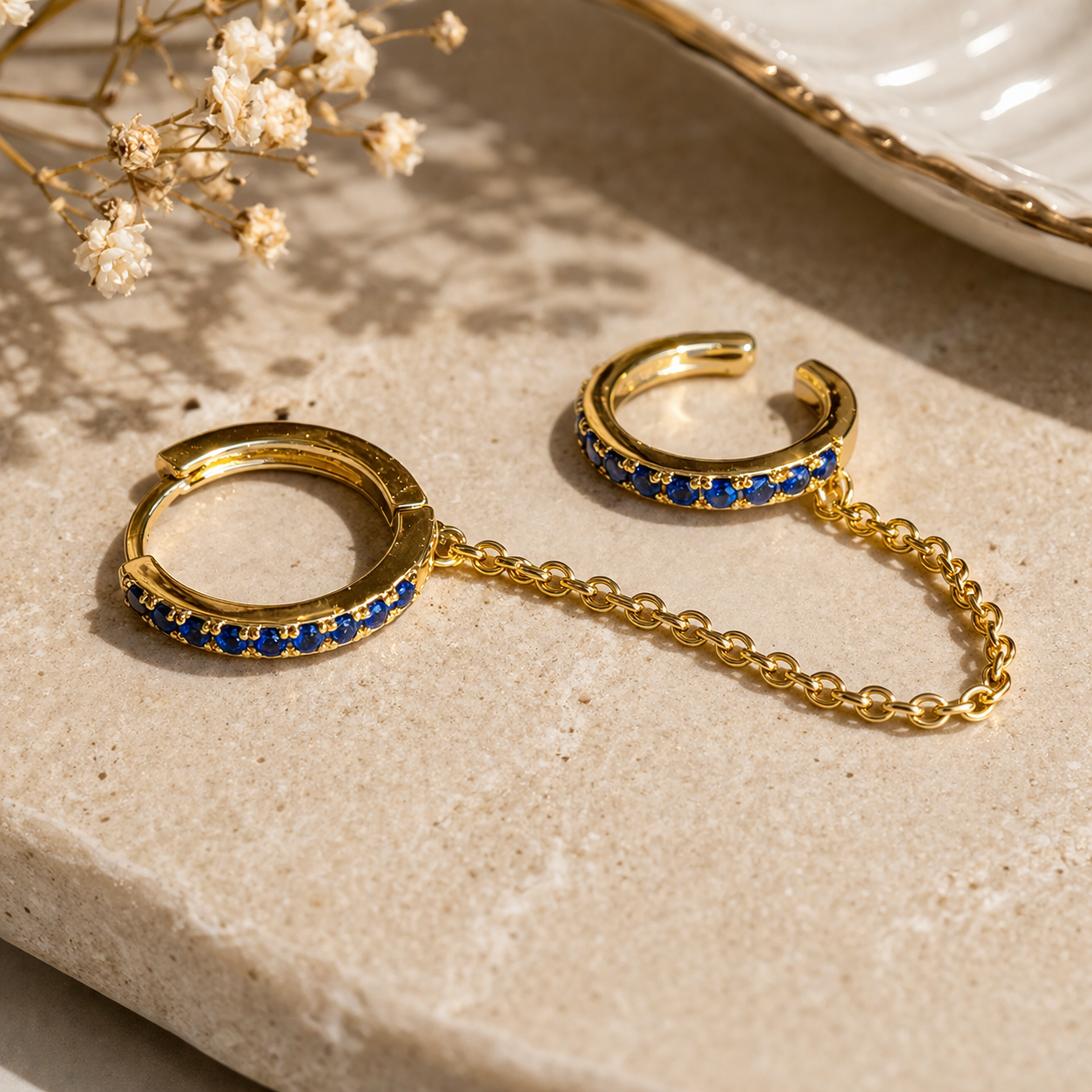 Gold hoop earrings with blue gemstones on a textured surface with dried flowers.