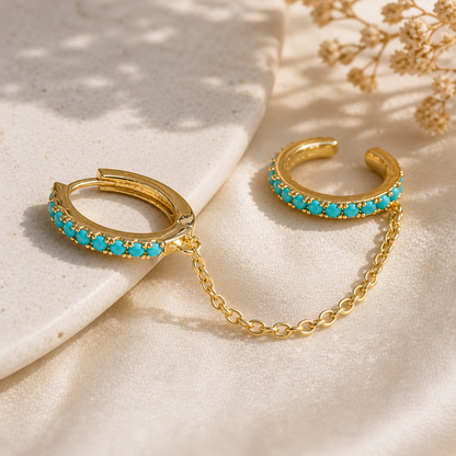 Gold hoop earrings with turquoise stones on a beige surface with dried flowers.