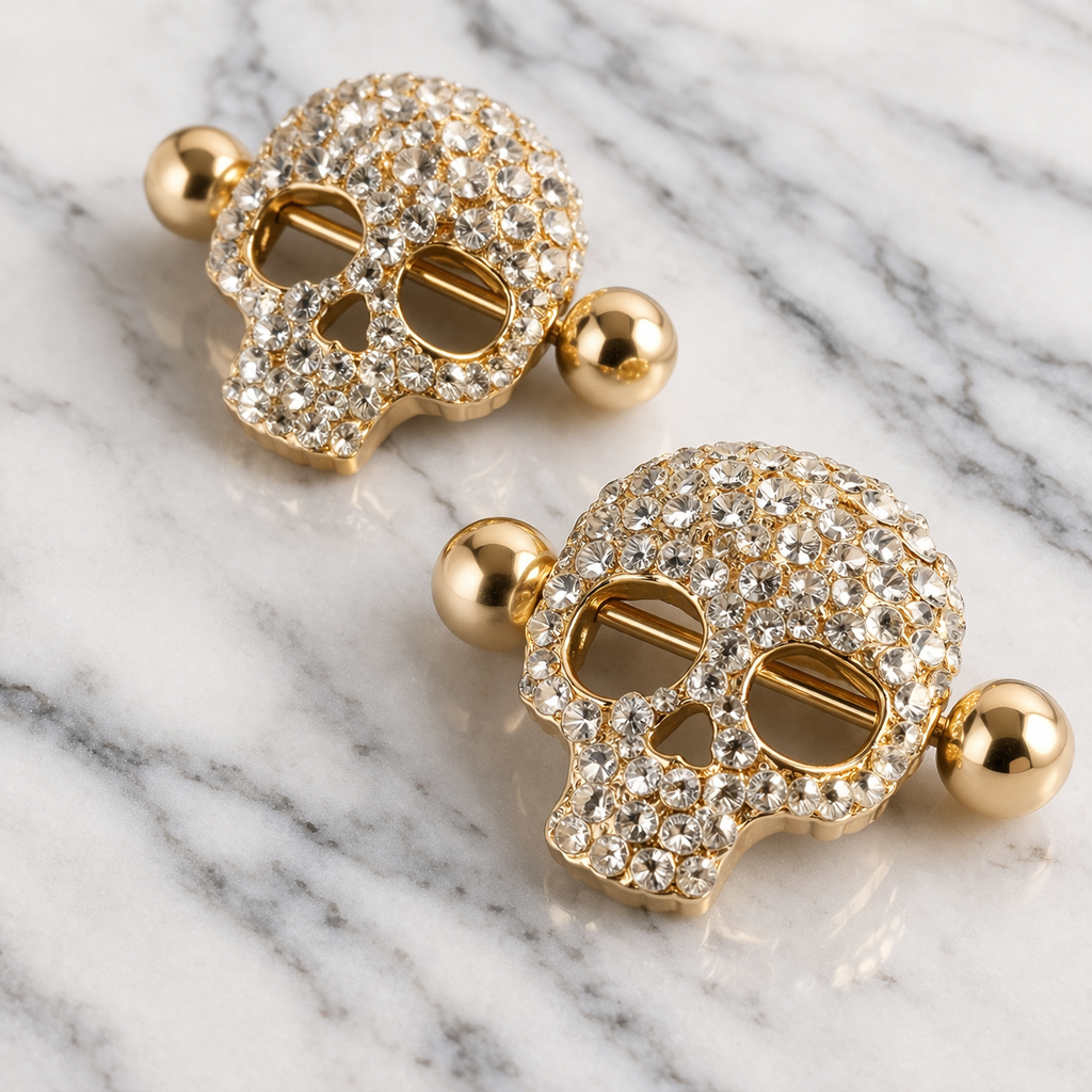 Gold skull-shaped nipple bars with crystal embellishments on a marble surface