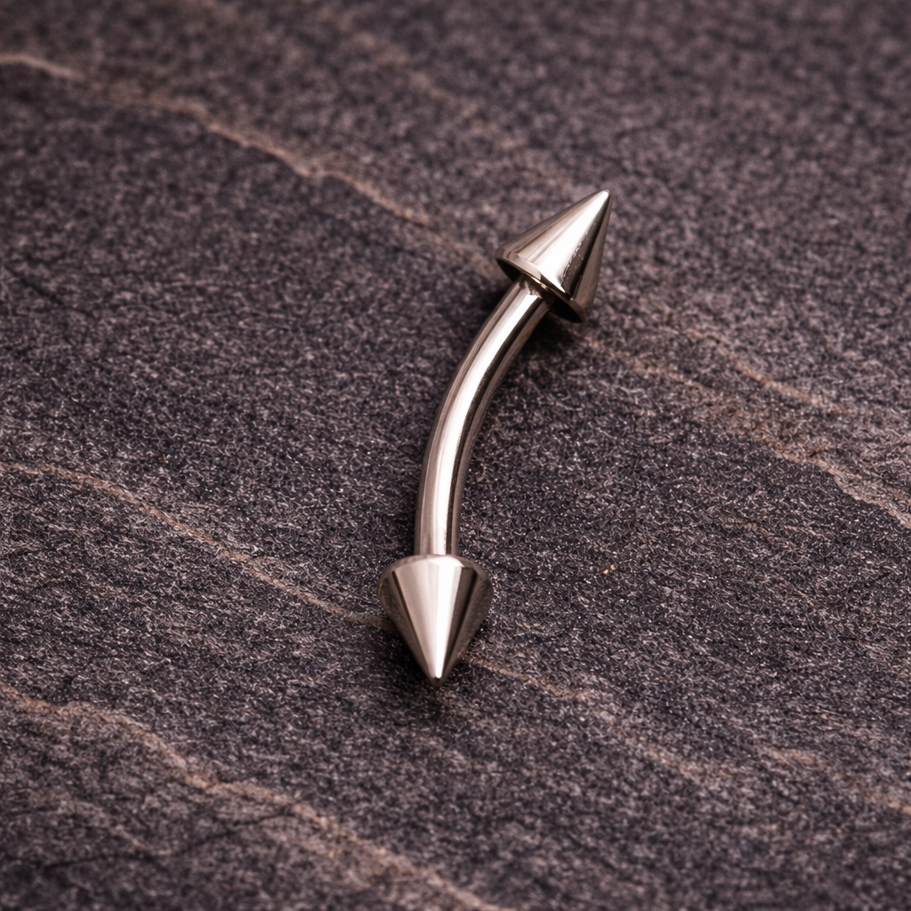 Metallic curved eyebrow/rook bar with cone-shaped ends on a textured brown surface