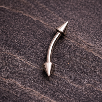 Metallic curved eyebrow/rook bar with cone-shaped ends on a textured brown surface