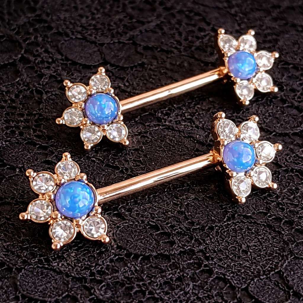 Pair of gold nipple bars with blue gemstones and clear stones on a black textured background