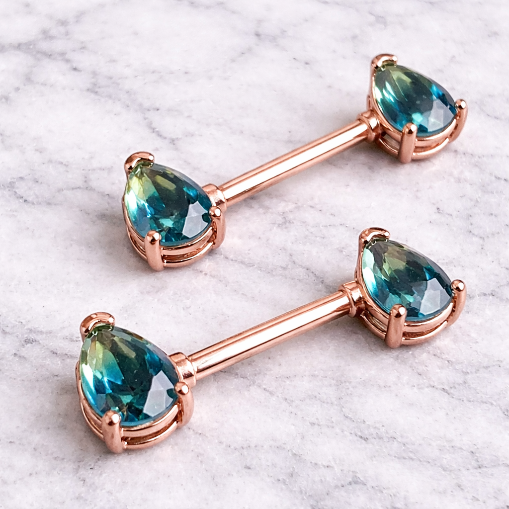 Pair of rose gold barbell nipple bars with blue gemstones on a marble background