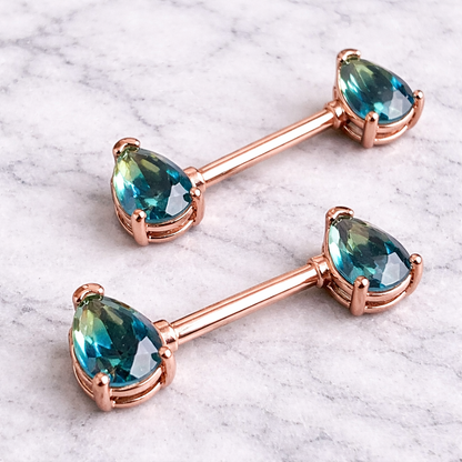Pair of rose gold barbell nipple bars with blue gemstones on a marble background