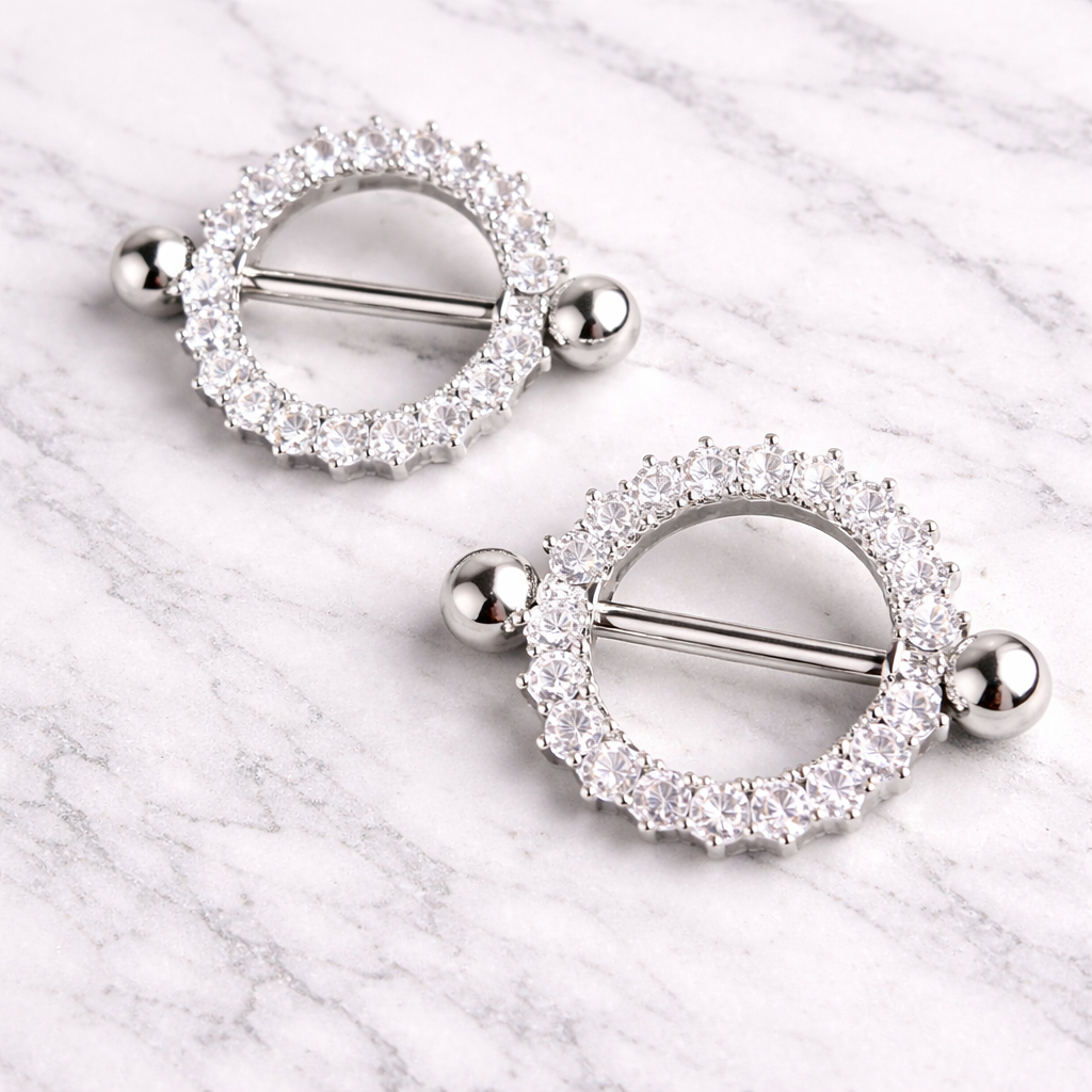 Pair of silver circular nipple bars with clear gemstones on a marble surface