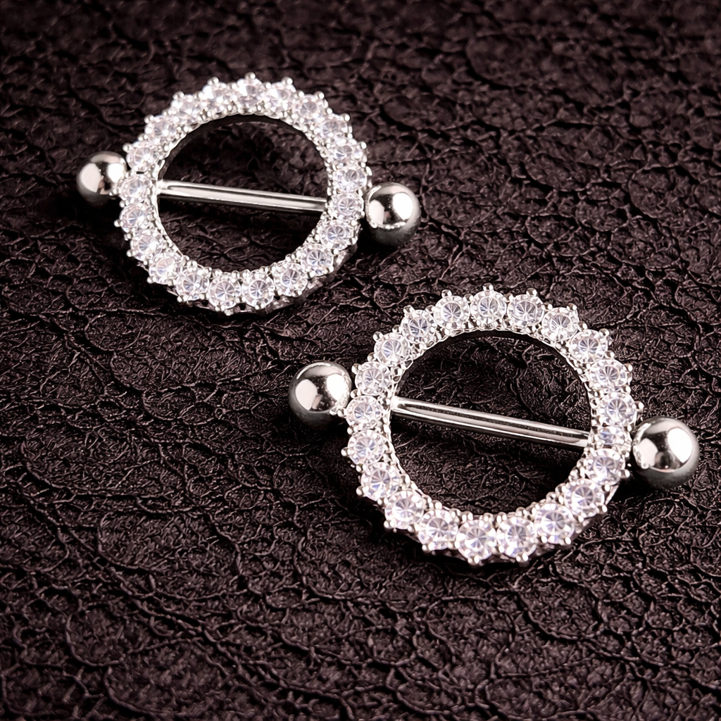 Pair of silver circular nipple shields with clear gemstones on a dark textured background