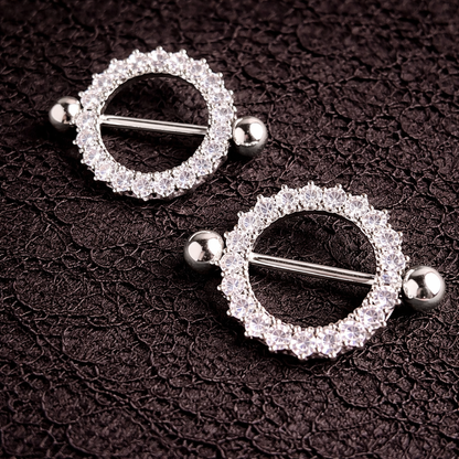 Pair of silver circular nipple shields with clear gemstones on a dark textured background