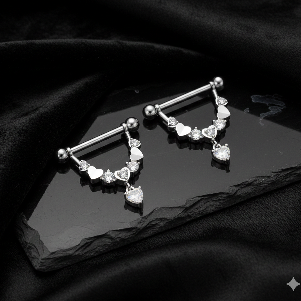 Pair of silver nipple barbells with heart and clear gemstone designs on a black surface.