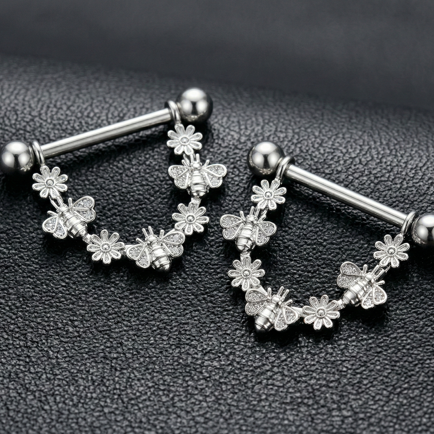 Pair of silver nipple piercing bars with floral designs on a black textured background