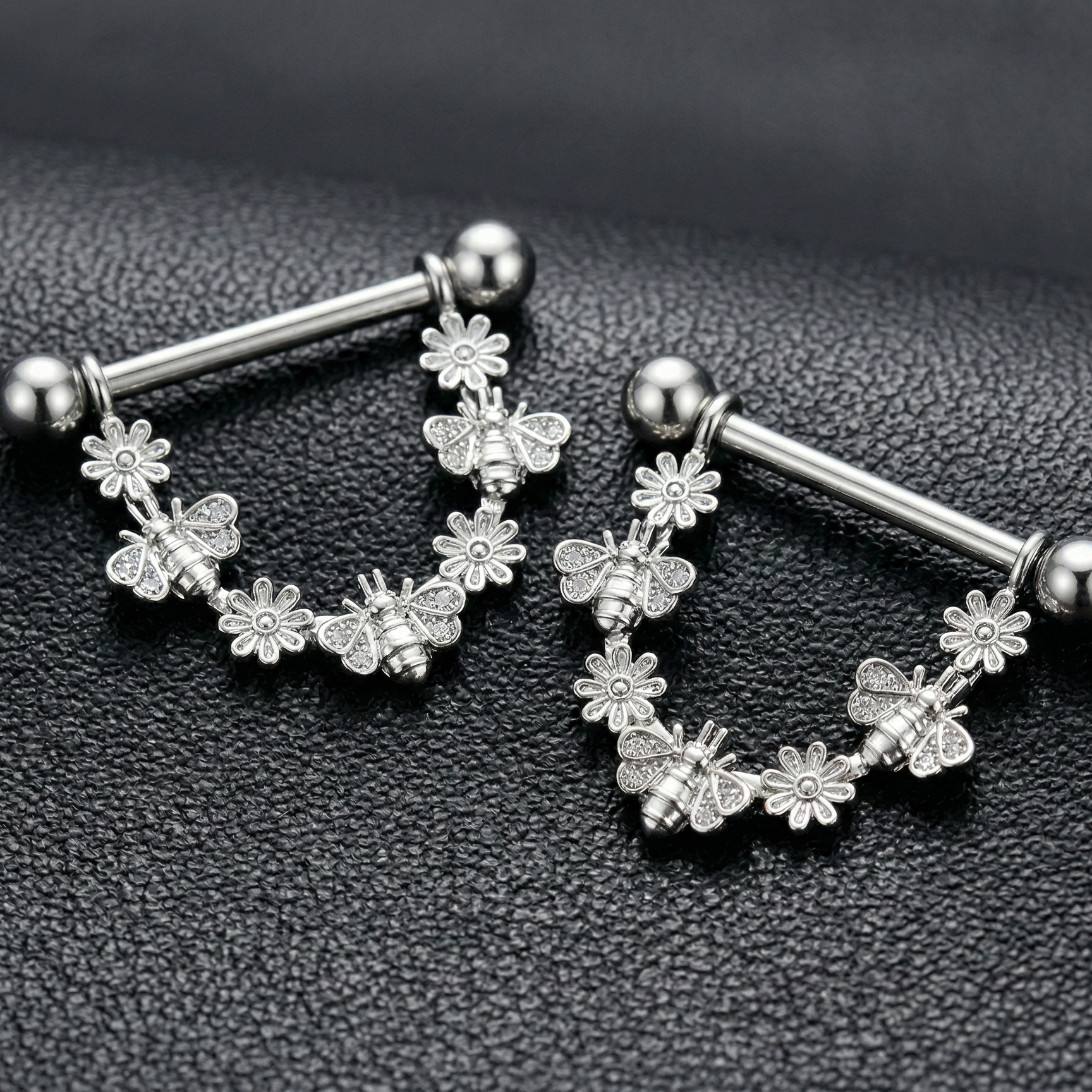 Pair of silver nipple piercing bars with floral designs on a black textured background