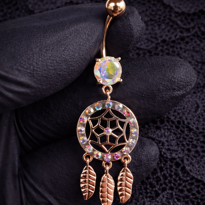 Navel ring with dreamcatcher design held by a gloved hand on a dark textured background