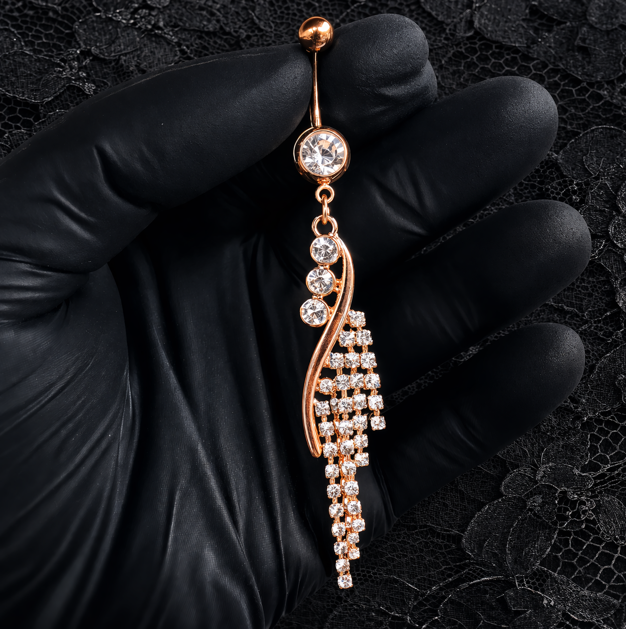 Rose gold navel bar with clear stones held by a gloved hand on a black lace background