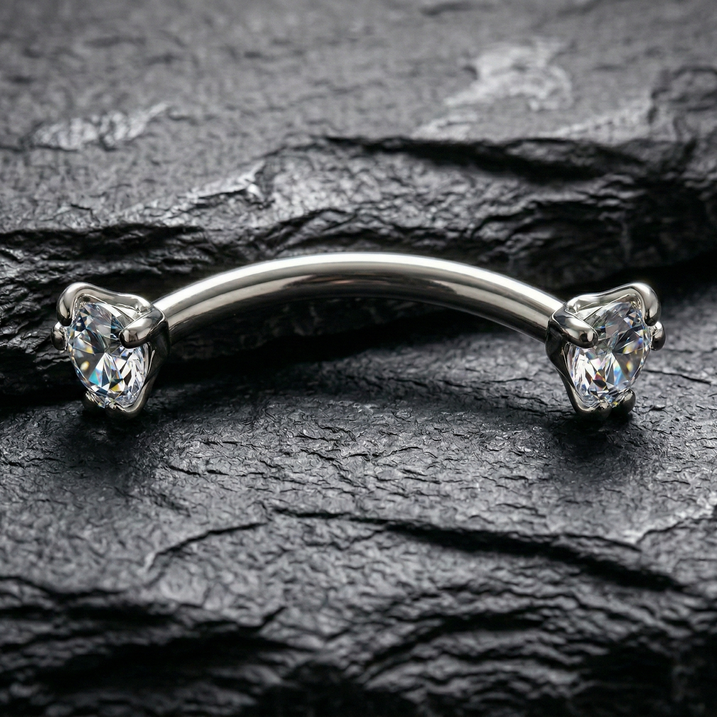 Silver eyebrow/rook bar with clear gemstones on a dark stone surface