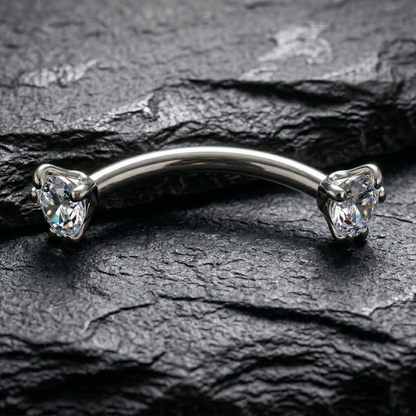 Silver eyebrow/rook bar with clear gemstones on a dark stone surface
