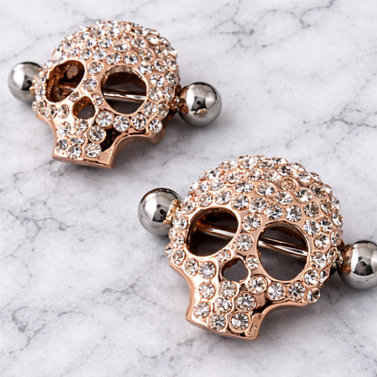 Two decorative skull nipple piercing bars with rhinestones on a marble surface