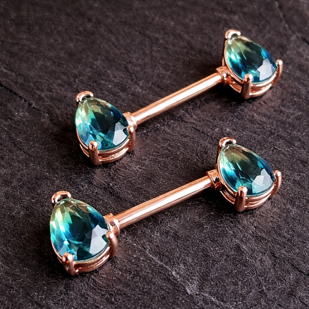 Two rose gold nipple barbells with blue teardrop stones on a dark surface