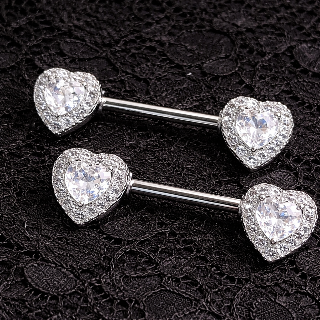 Two silver nipple barbells with heart-shaped ends featuring clear gemstones on a black textured background.