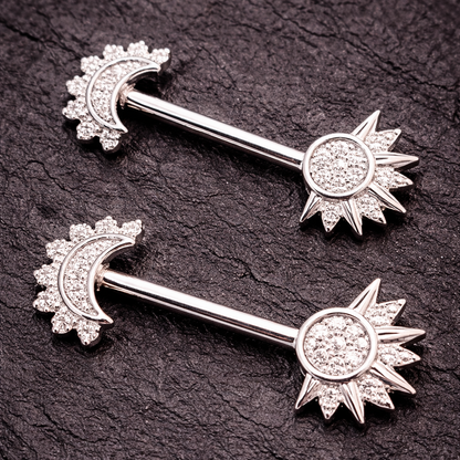 Two silver nipple bars with sun and moon designs on a dark textured background